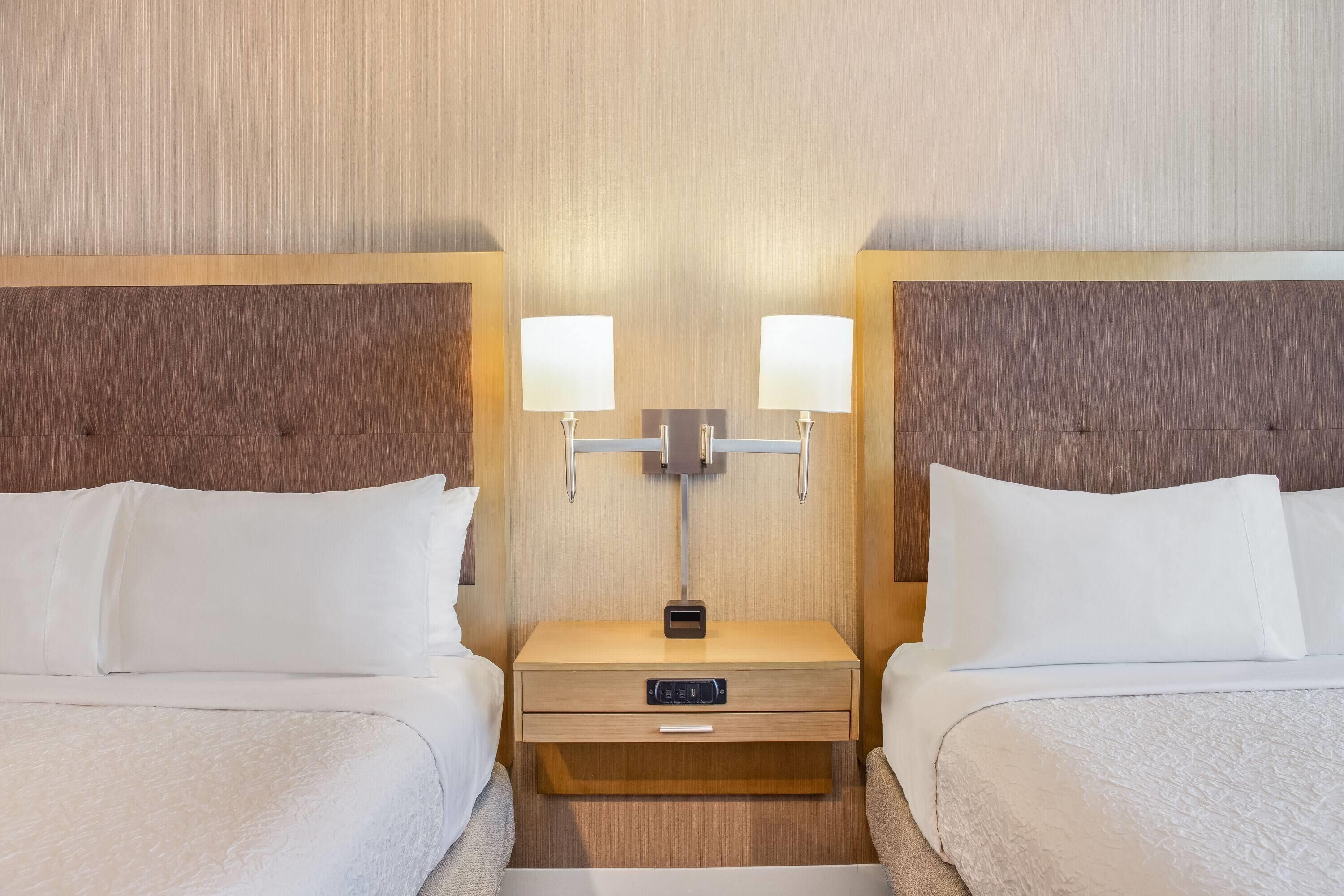 A hotel room with two beds, a bedside table with a lamp, and a control panel.
