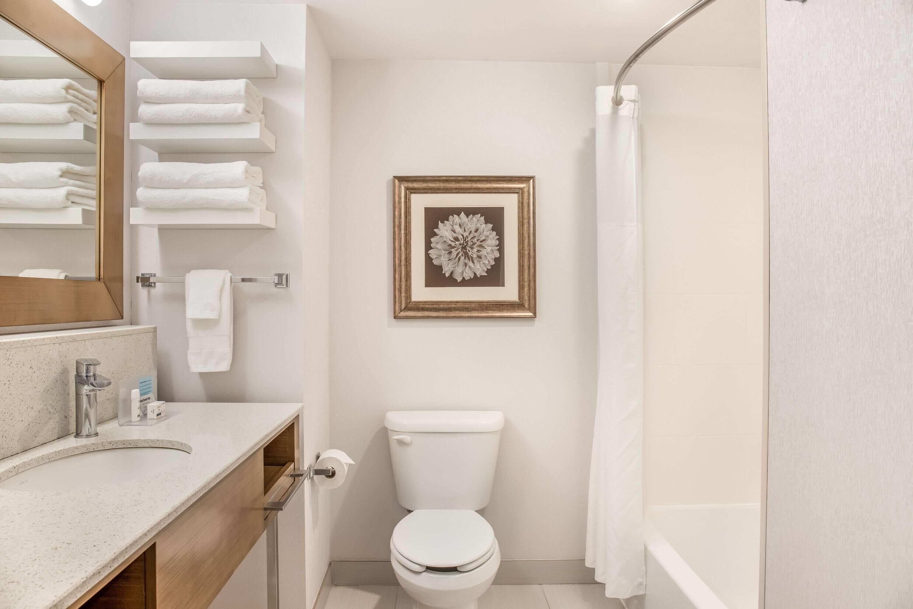 Een moderne badkamer met toilet, wastafel en douche. Handdoeken liggen op een rek en er hangt een afbeelding in een lijst aan de muur.