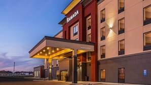 Une entrée d’hôtel moderne, avec un passage couvert, une enseigne indiquant « RAMADA » et un panneau d’accueil.