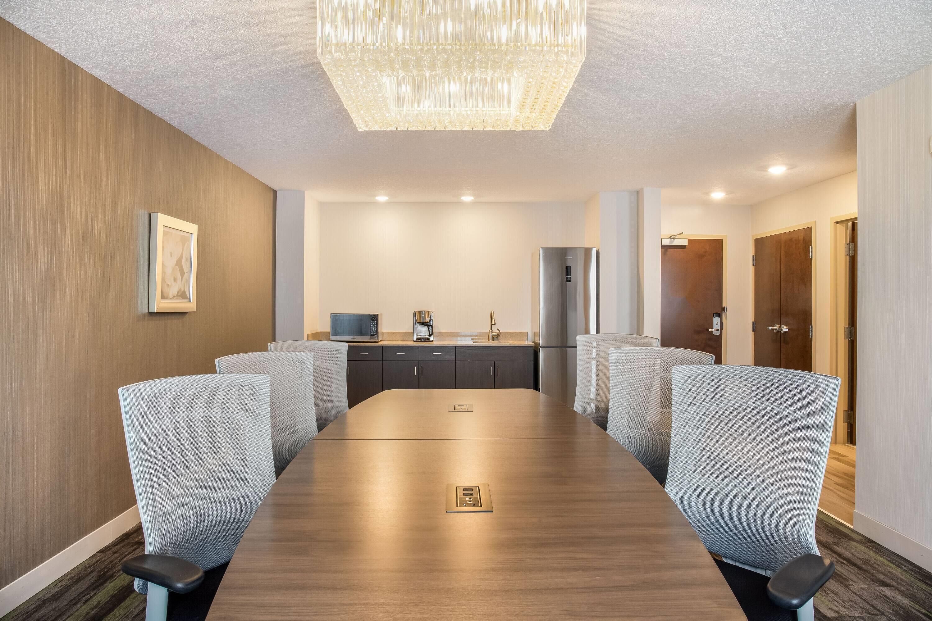 A conference room with a long table, six chairs, a chandelier, a refrigerator, and a microwave in the background.