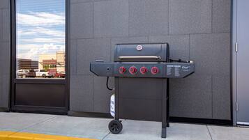A portable grill with four burners and a digital display, positioned on a sidewalk next to a building with a large window.