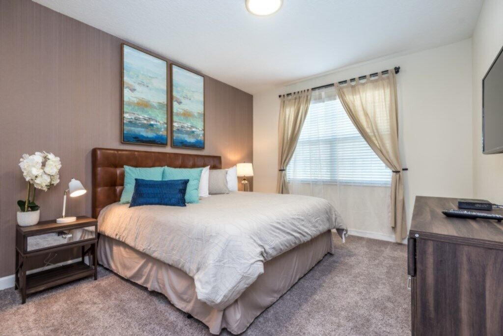 Ein Hotelzimmer mit einem großen Bett, einem Nachttisch mit Lampe, einem Schreibtisch mit Computer und einem Fenster mit Vorhängen.