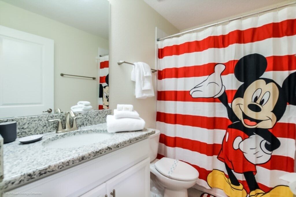 Un baño con cortina de ducha de Mickey Mouse, un inodoro blanco y una encimera de granito con lavabo y toallero.