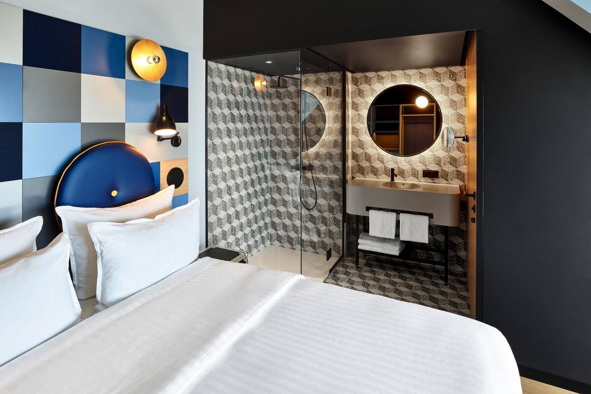 Une chambre d’hôtel moderne avec un grand lit, une salle de bain dotée d’un grand miroir rond et un motif à carreaux sur le mur.