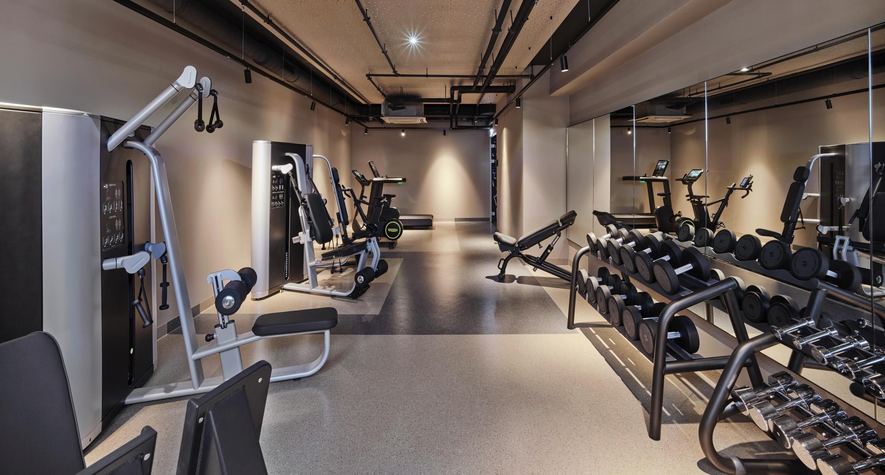 Et fitnesscenter med forskellig træningsudstyr, herunder løbebånd, crosstrainere og en vægtstang med håndvægte.