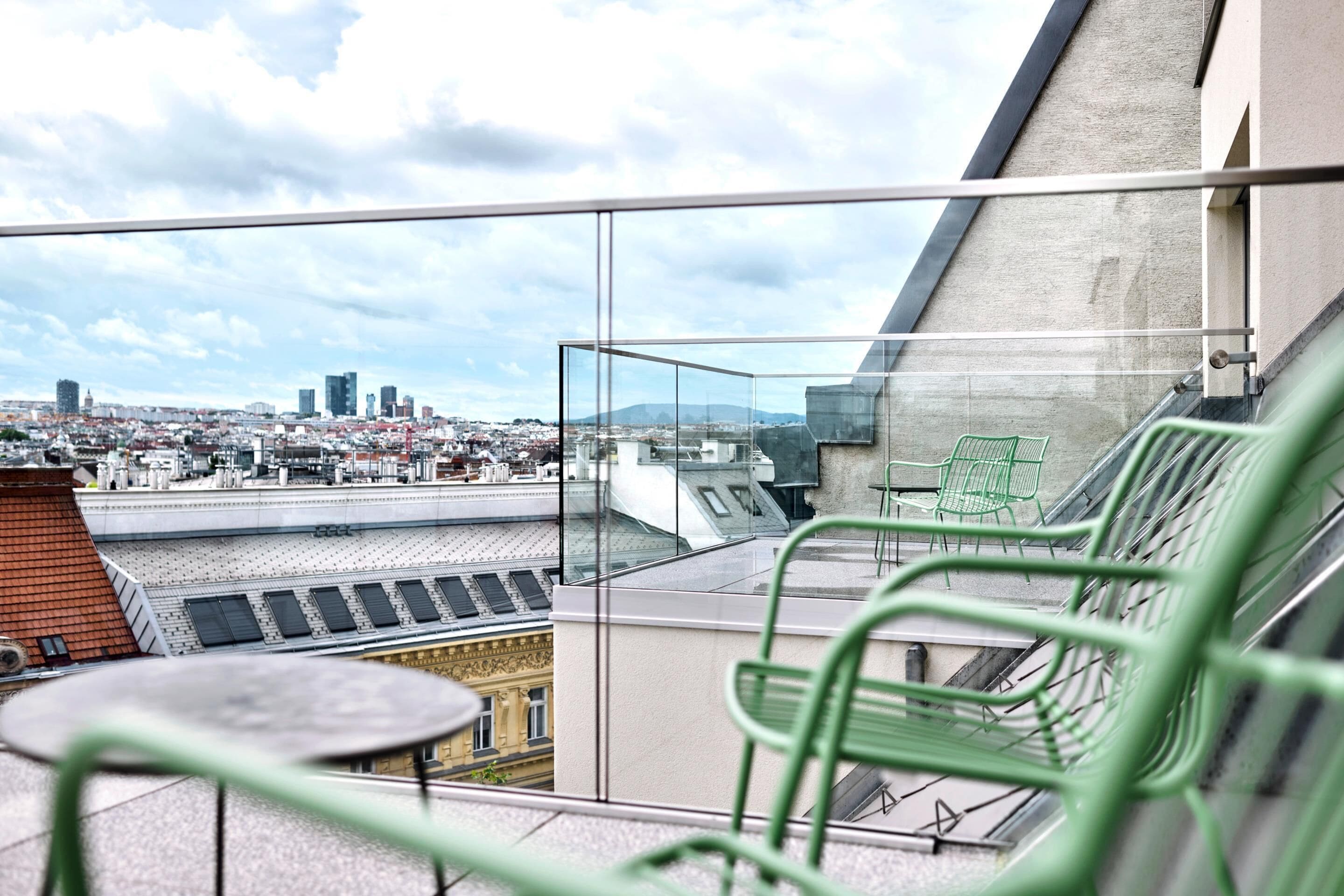 Un balcone con ringhiera in vetro, un tavolo e una sedia, con vista su un paesaggio urbano che include edifici e una montagna all'orizzonte.