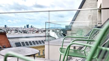 Un balcone con ringhiera in vetro, un tavolo e una sedia, con vista su un paesaggio urbano che include edifici e una montagna all'orizzonte.