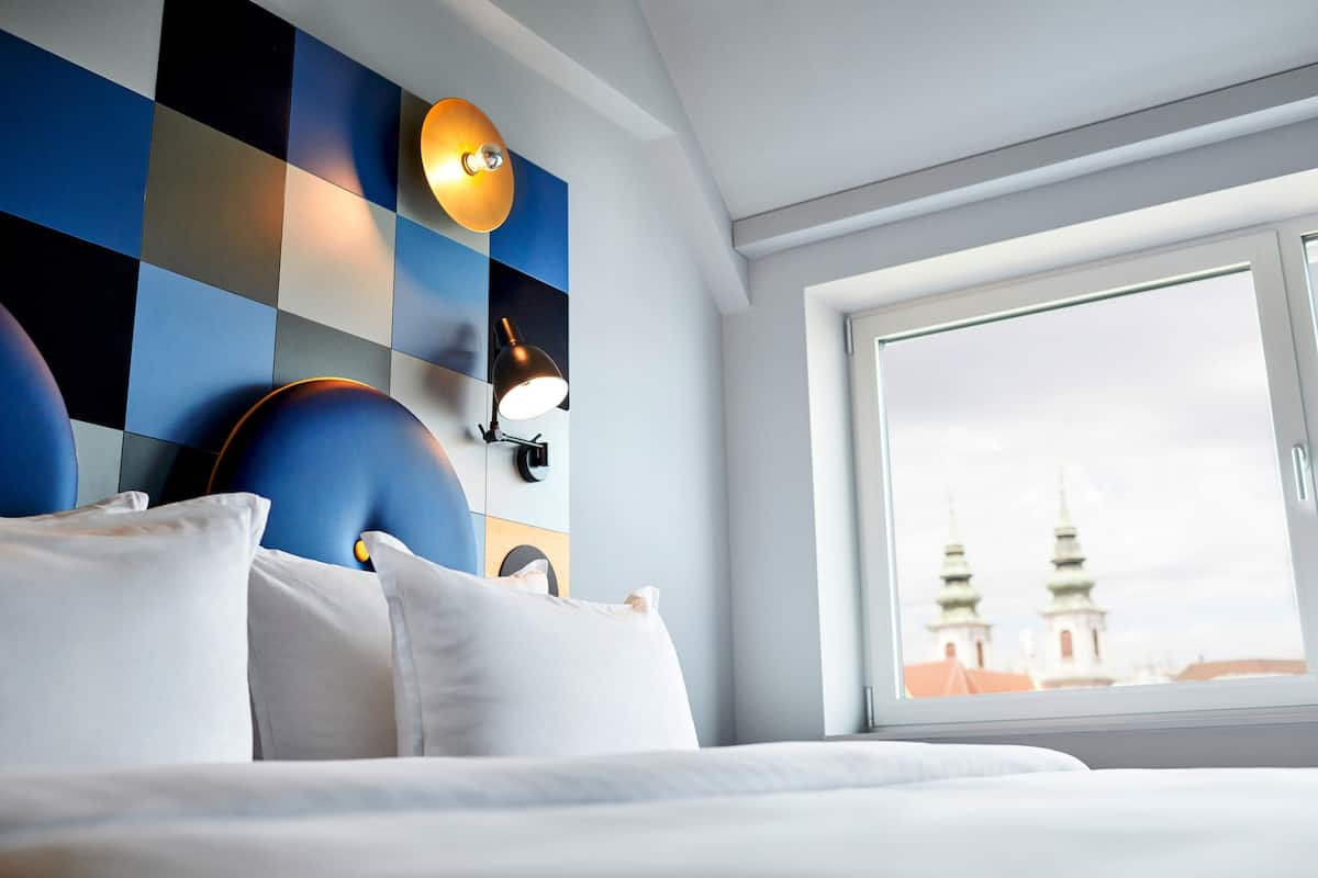 Une chambre d’hôtel avec un mur à carreaux, un lit avec du linge de lit blanc et une fenêtre donnant sur des immeubles.