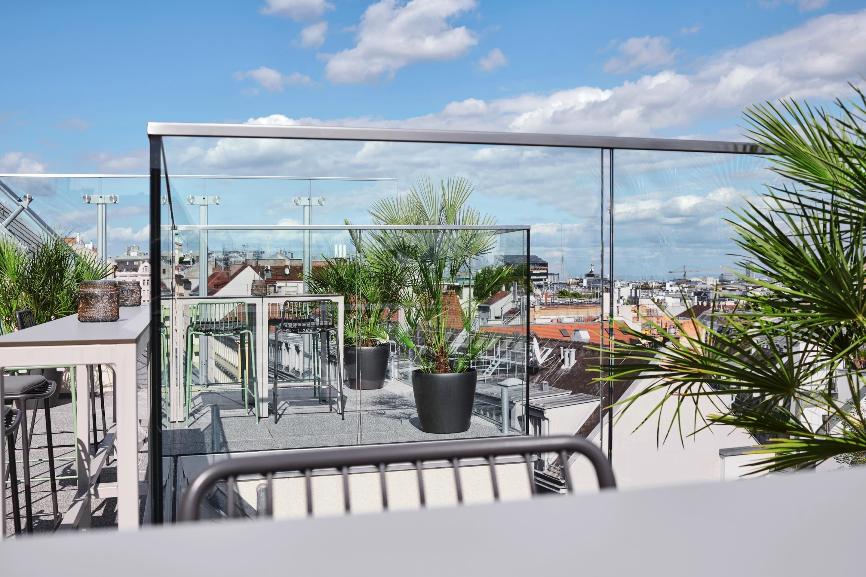 Terraço no telhado com plantas em vasos, guarda-corpo de vidro e vista para a paisagem urbana.