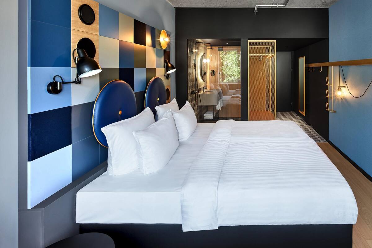Une chambre d’hôtel moderne, dotée d’un grand lit, d’un coin salon et d’une salle de bain visible à travers une porte ouverte.