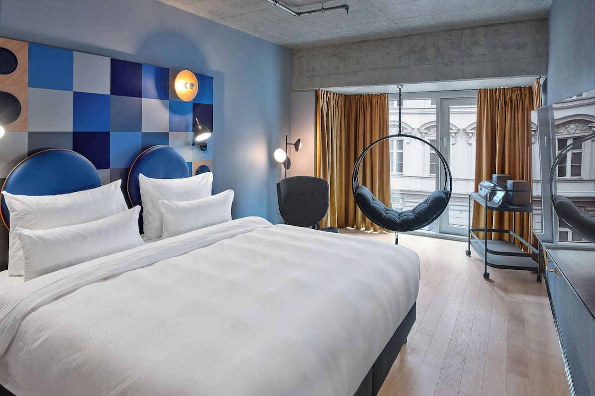 Une chambre d’hôtel moderne dotée d’un grand lit, d’un bureau avec une chaise, d’un fauteuil suspendu et offrant une vue sur la ville grâce à de grandes fenêtres.