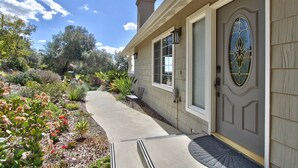 A house with a well-maintained garden, a front door with a decorative glass panel, and a visible chimney. - Hilltop Comforts (Fallbrook)