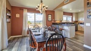 A dining area with a wooden table set for six, surrounded by chairs, a chandelier, and a view of the outdoors. - Hilltop Comforts (Fallbrook)
