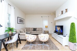 A living room with a sofa, two patterned chairs, a dining table, a TV, and a potted plant.
