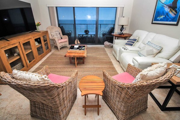 A living room with a television, wicker furniture, a white sofa, and a view of the ocean through large windows. - Direct Oceanfront Views from Luxury Condo in Southwind Complex! (New Smyrna Beach)
