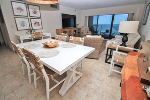 A spacious living area with a dining table, wicker furniture, a TV, and a balcony with a view. - Direct Oceanfront Views from Luxury Condo in Southwind Complex! (New Smyrna Beach)