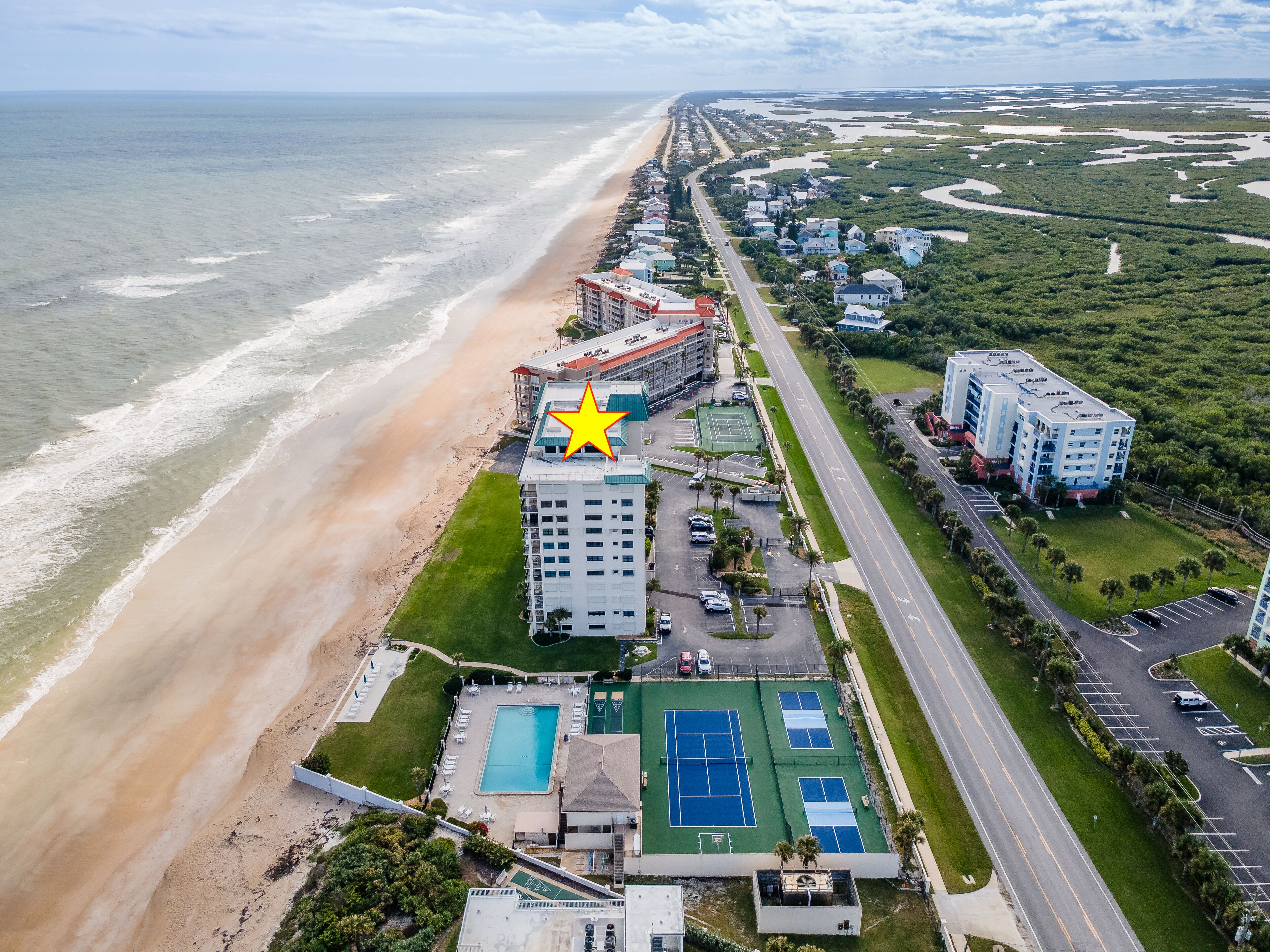 Luftaufnahme eines Hotels an der Küste mit Strand, Tennisplätzen und einem markierten Gebäude.