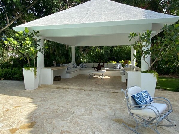 A covered outdoor seating area with stone flooring, white furniture, and potted plants. - Lux Villa, Casa de Campo (La Romana)