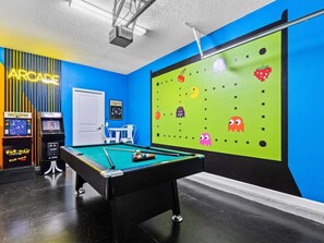 A pool table with a green felt surface, two Pac-Man arcade machines, and a wall with a green screen displaying pixelated ghosts. - 6BR Solterra Pool Home | Jacuzzi, Game Room, BBQ & 2 King Suites (Davenport)