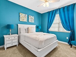 A bedroom with a white bed, two nightstands, a ceiling fan, and a window with blue curtains.