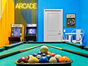A pool table with balls, two Pac-Man arcade machines, a door, and a blue wall with a neon sign.
