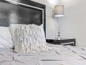 Una cama con un cabecero texturizado, una lámpara sobre una mesita de noche y una pared de color neutro.