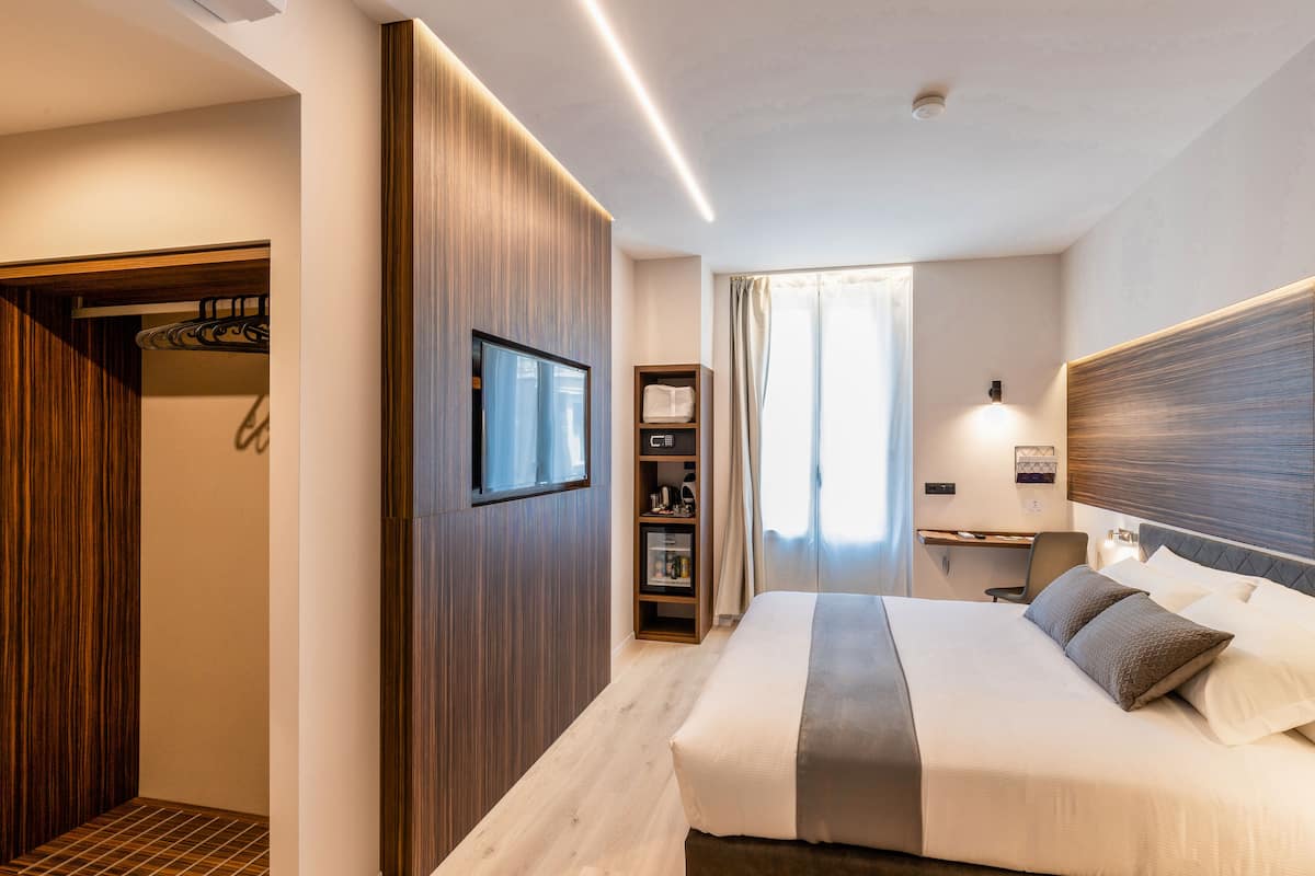 A modern hotel room with a large bed, a desk with a chair, a wardrobe, and a television.
