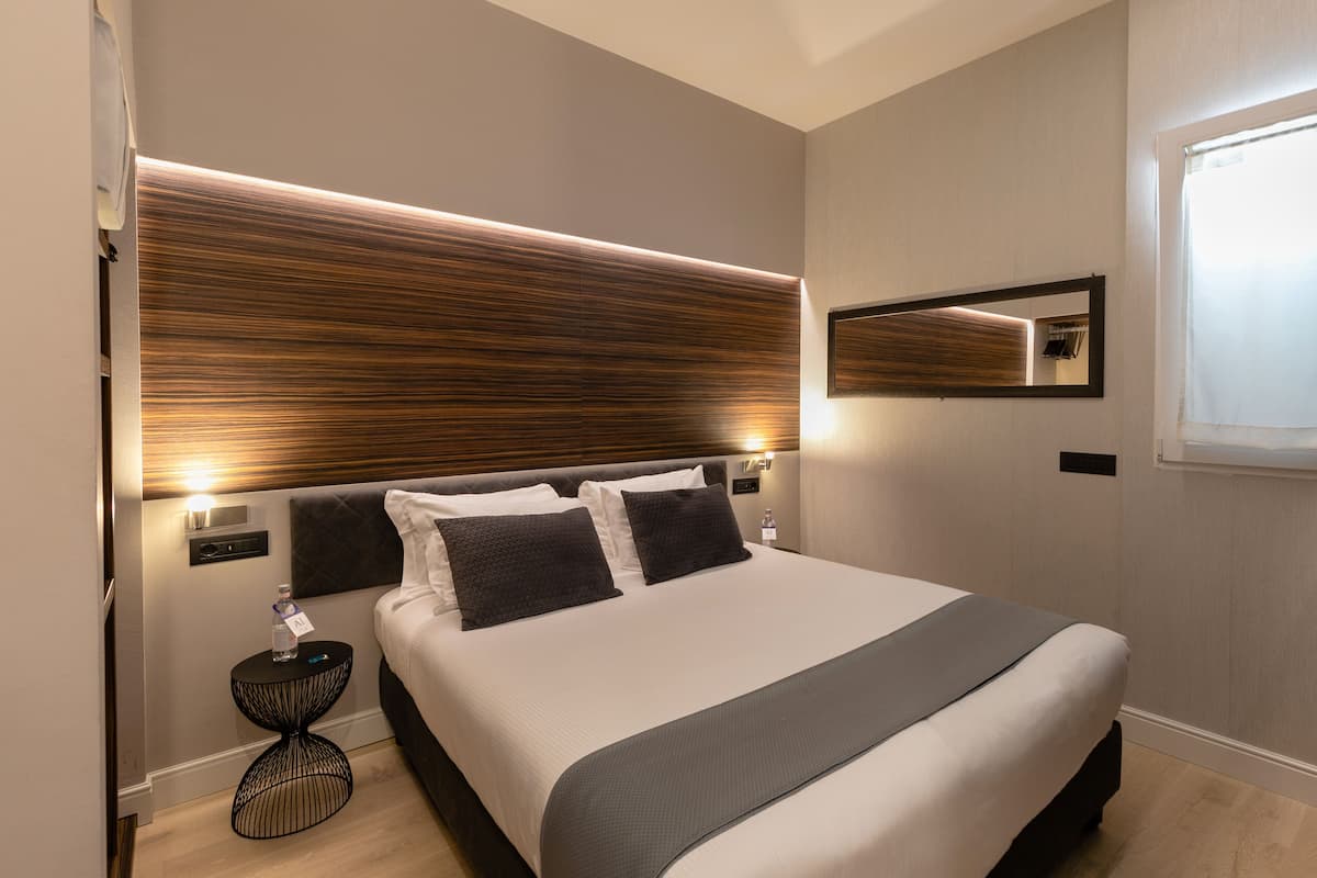 A modern hotel room with a large bed, a bedside table with a bottle, a mirror, and a window with curtains.