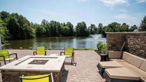 Une terrasse extérieure avec une table en pierre, des chaises jaunes et un canapé beige donnant sur un lac agrémenté d’une fontaine.