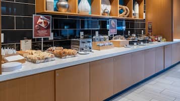 Un bufé de desayuno con diversos pasteles y panes, una máquina de café y un cartel anunciando bollería para el desayuno.