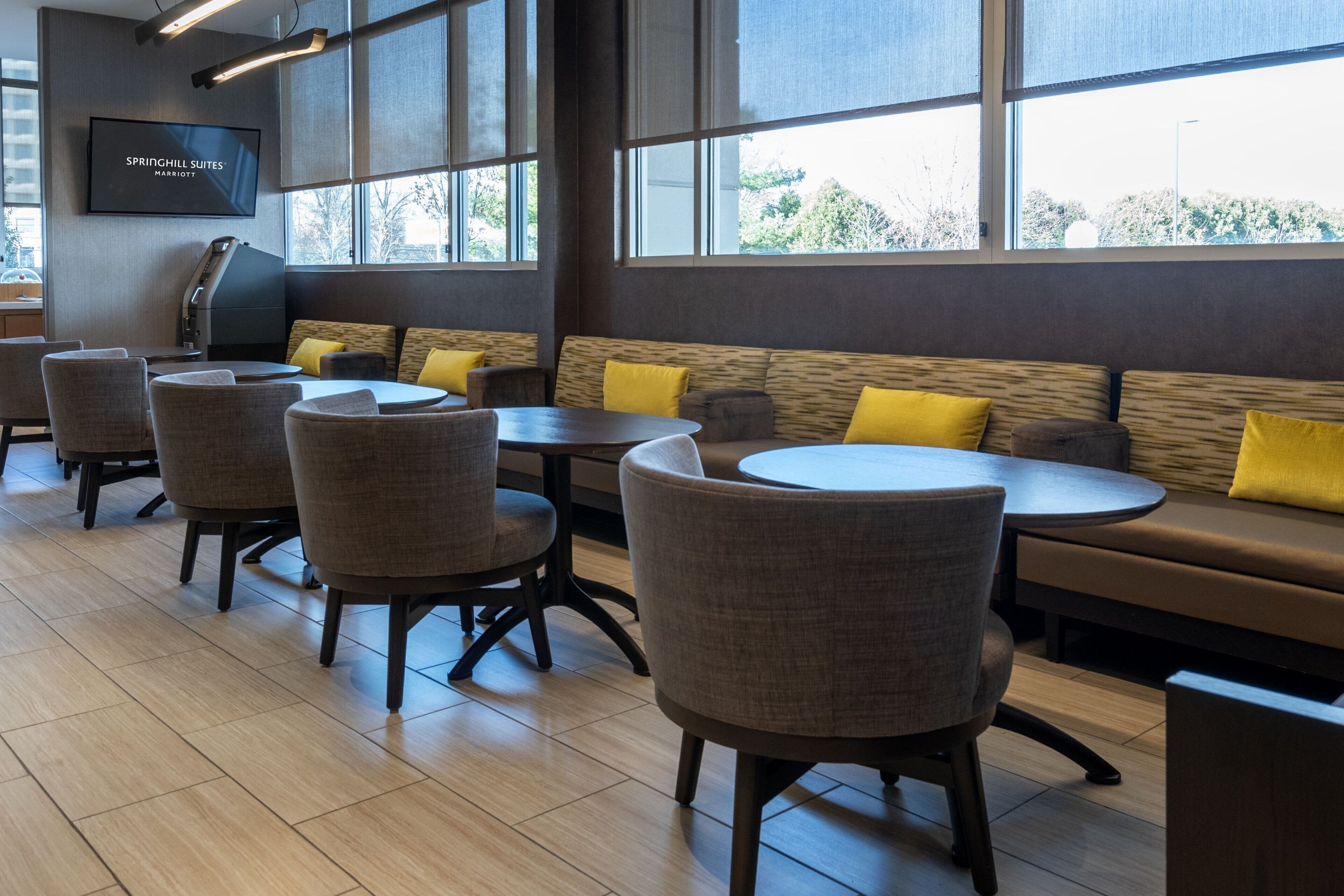 A modern restaurant with a seating area featuring round tables and cushioned chairs, a large window with a view, and a flat-screen TV mounted on the wall.