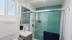 Et badeværelse med et hvidt toilet, en håndvask, et spejl, en brusekabine med glasvægge og et blåt håndklæde hængende på væggen.