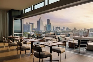 A modern rooftop lounge with a city view, featuring tables, chairs, and a sofa area.