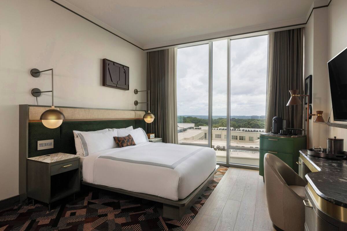 A hotel room with a large bed, a nightstand, a desk, a chair, a television, and a view of the city through large windows.