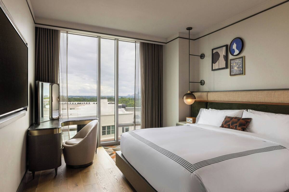 A hotel room with a large bed, a desk, a chair, and a view of the city through large windows.