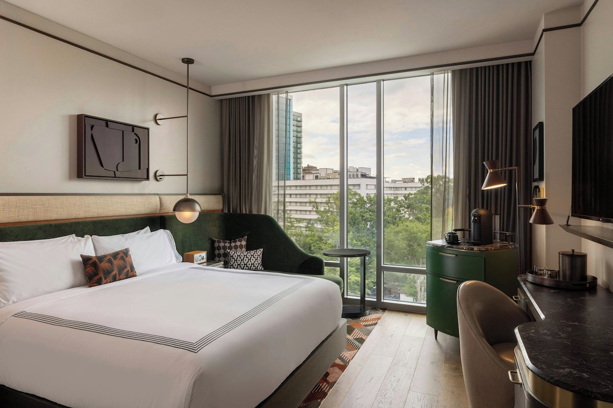 Una habitación de hotel con una cama grande, un sillón verde, una mesita redonda pequeña, un escritorio con lámpara y un ventanal con vistas a la ciudad.