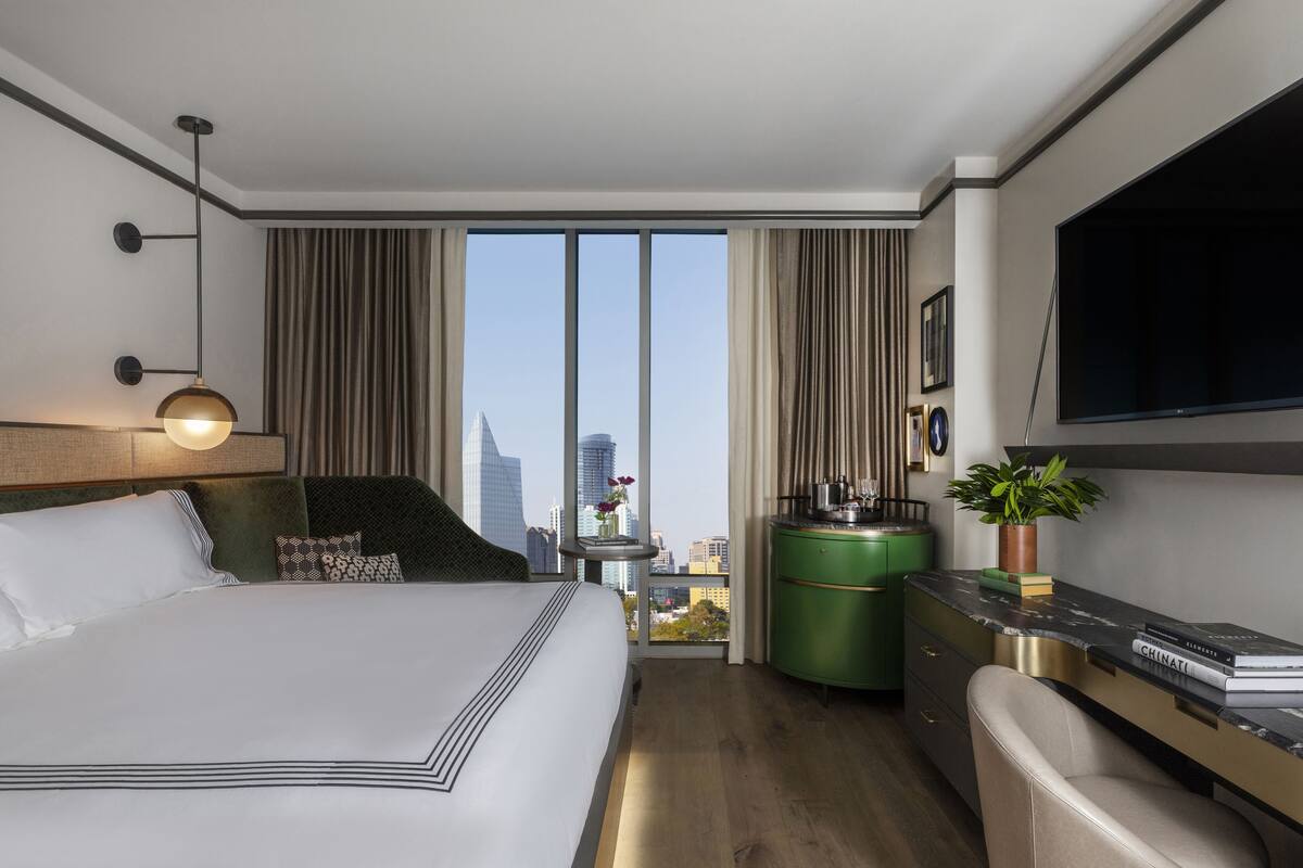 A modern hotel room with a large bed, a desk, a chair, a television, and a view of the cityscape.