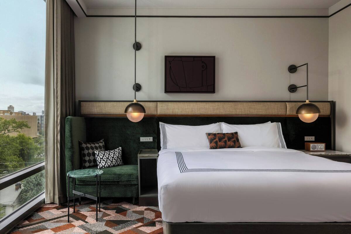 A hotel room with a large bed, a green armchair, a small table, and a view of the city.