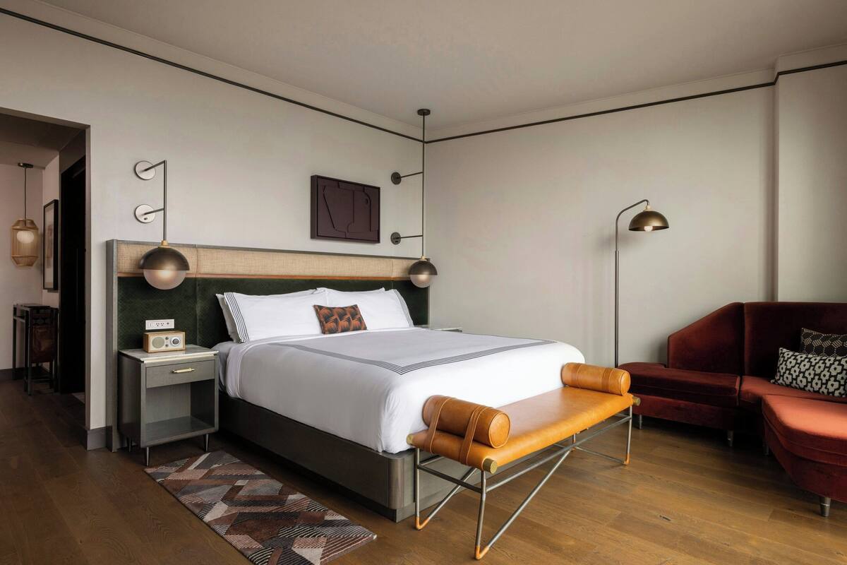 A modern hotel room with a large bed, a wooden headboard, a bedside table with a radio, a red sofa, and a patterned rug.