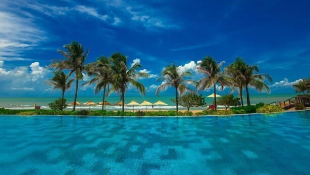 A swimming pool with clear blue water, surrounded by palm trees and beach umbrellas, with a view of the ocean and a clear sky. - InterContinental Grand Ho Tram (Ho Chi Minh City)