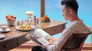 A man reading a magazine at a table with a view of the sea, a fruit bowl, and a vase of flowers. - InterContinental Grand Ho Tram (Ho Chi Minh City)