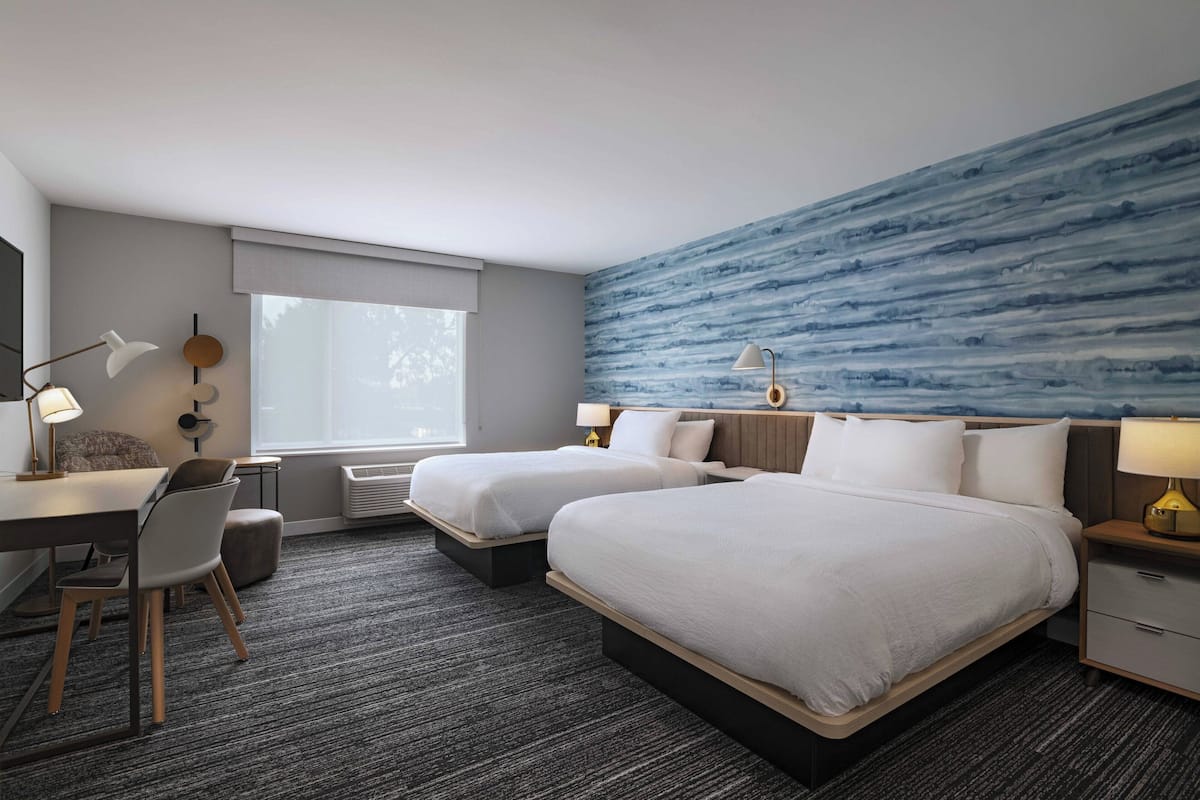 Habitación de hotel con dos camas, un escritorio, una silla, una lámpara y un ventanal grande.