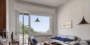 A modern living room with a sofa, coffee table, and dining table. A large window offers a view of a cityscape.