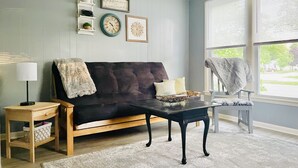 Una sala de estar con un sofá marrón oscuro, una mesa de centro de madera, una mesita auxiliar con una lámpara y un ventanal con persianas.