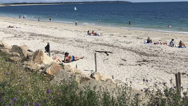 Ein Strand mit Menschen, die sonnenbaden und schwimmen, ein Segelboot auf dem Wasser und ein klarer Himmel.