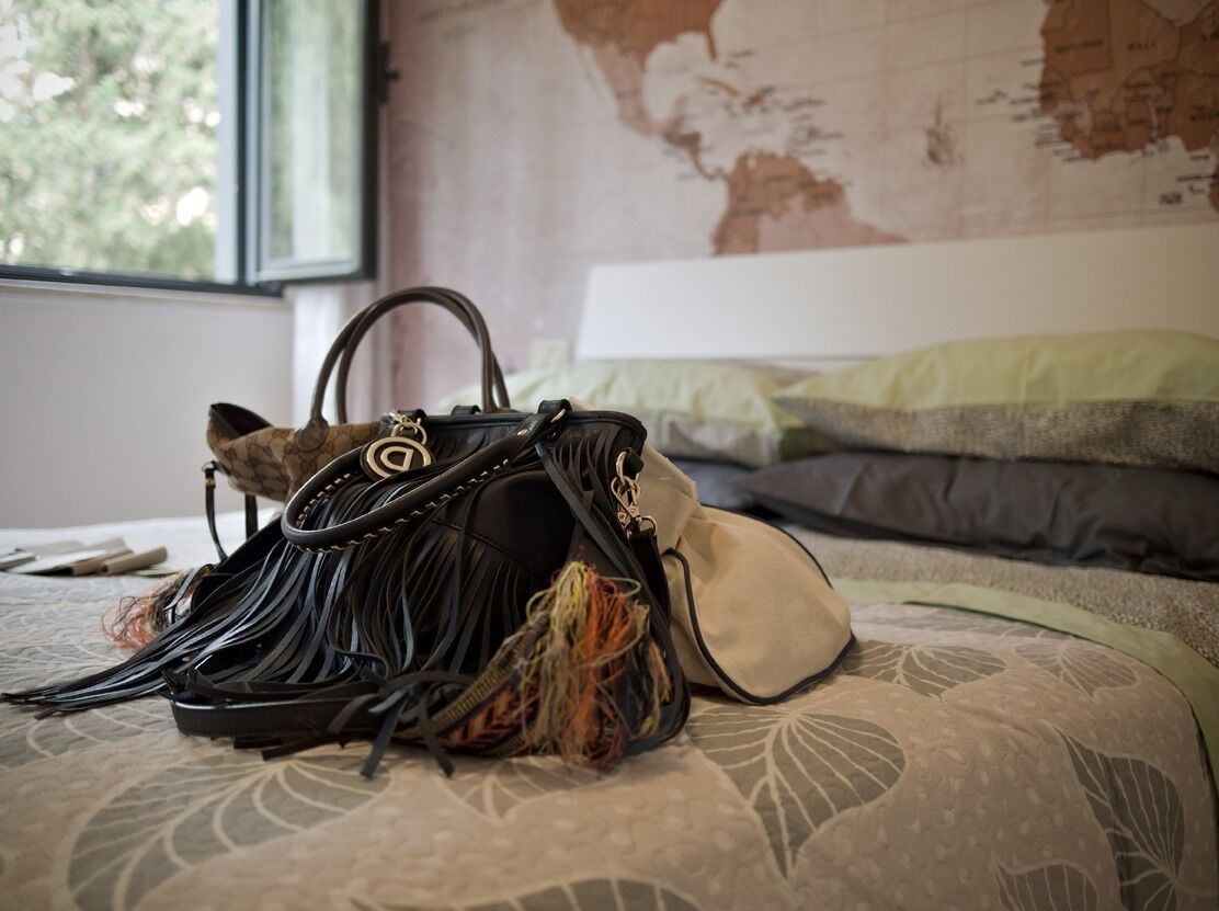A bed with a patterned bedspread and pillows, a window with a view of trees, and a black handbag with tassels on the bed.