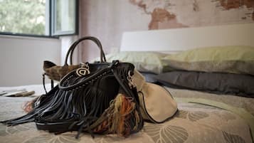 A bed with a patterned bedspread and pillows, a window with a view of trees, and a black handbag with tassels on the bed.