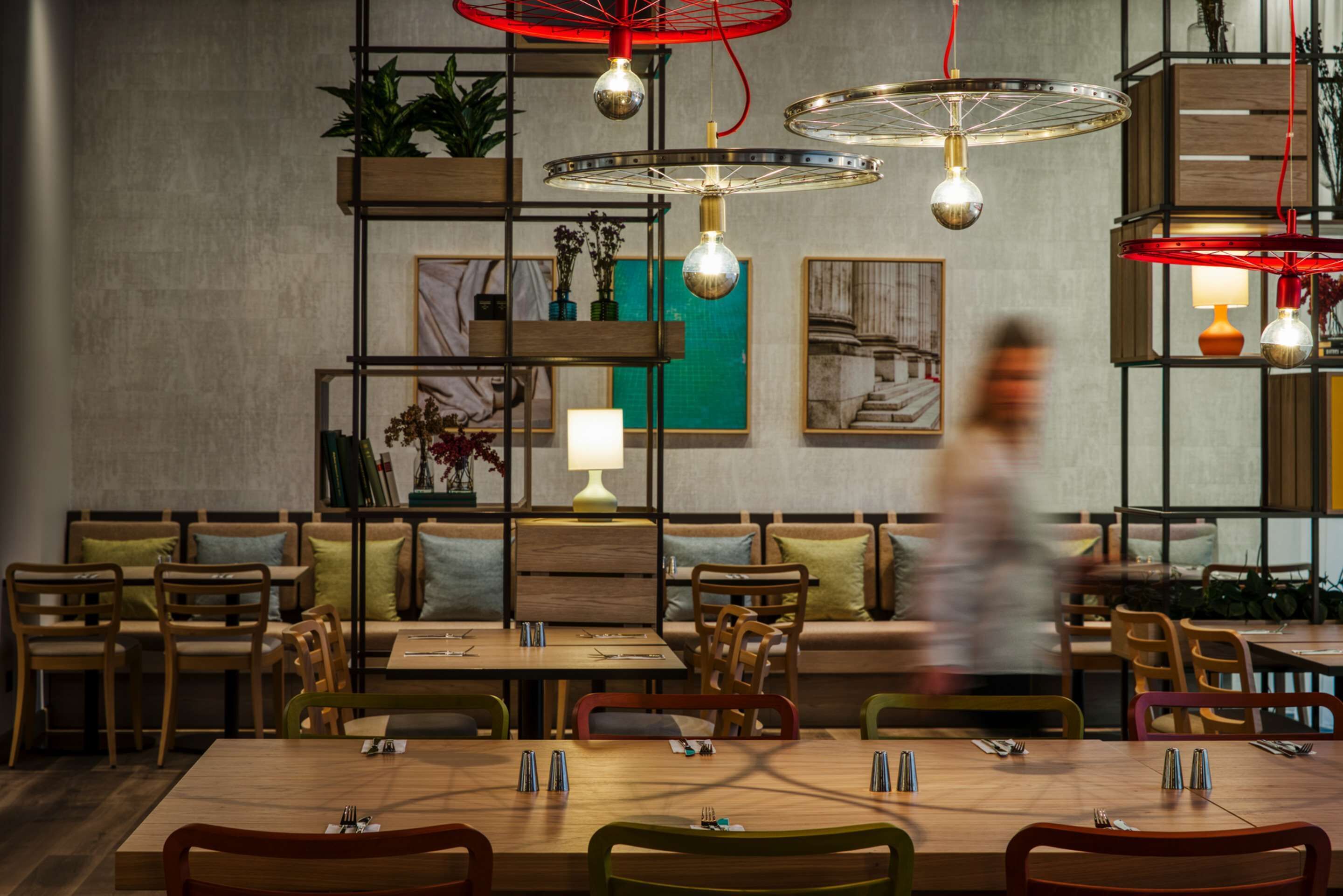 A restaurant interior with wooden tables and chairs, hanging pendant lights, and shelves with plants.