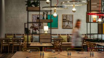 A restaurant interior with wooden tables and chairs, hanging pendant lights, and shelves with plants.