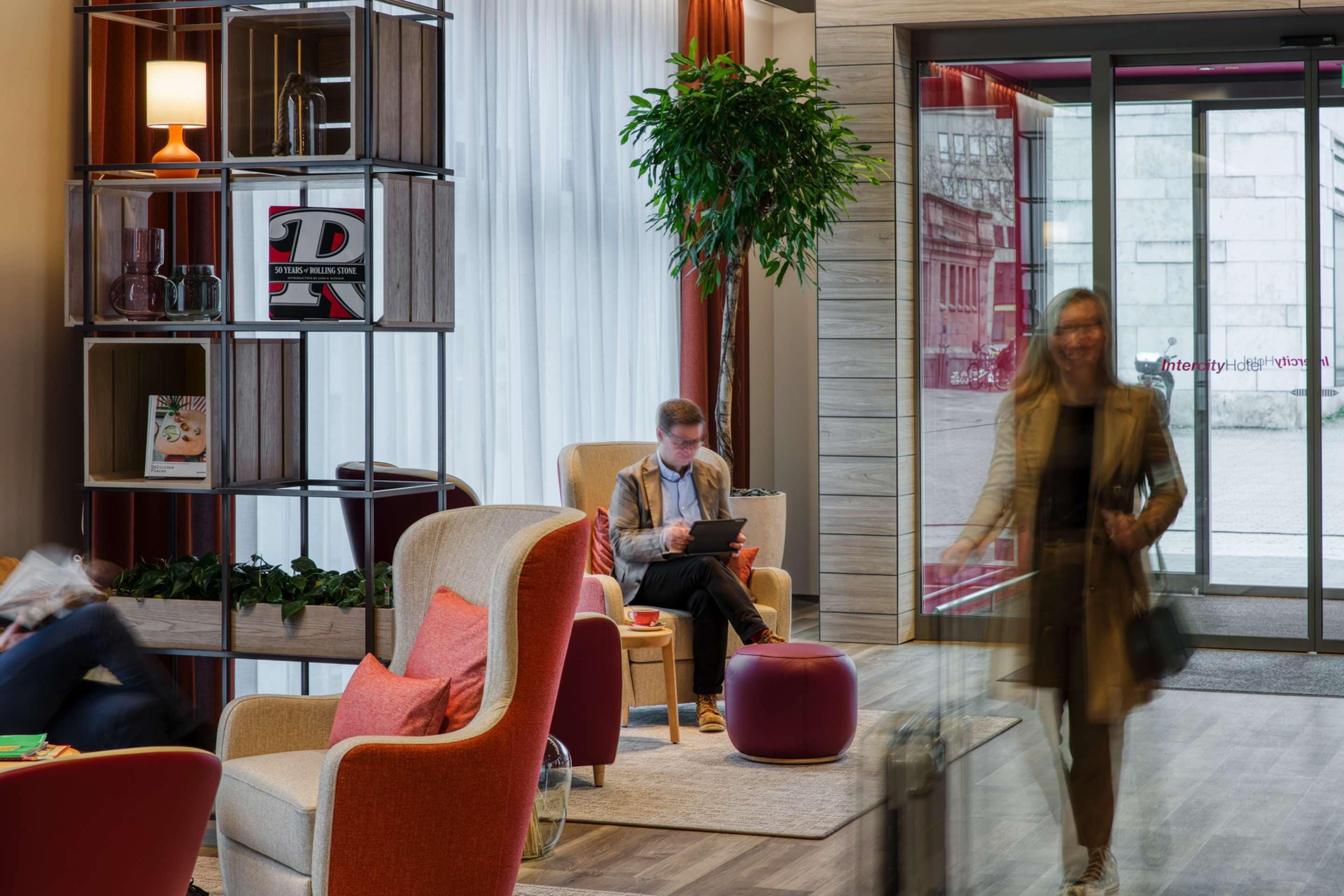 A modern hotel lobby with a seating area, a coffee machine, a bookshelf, and a large window.